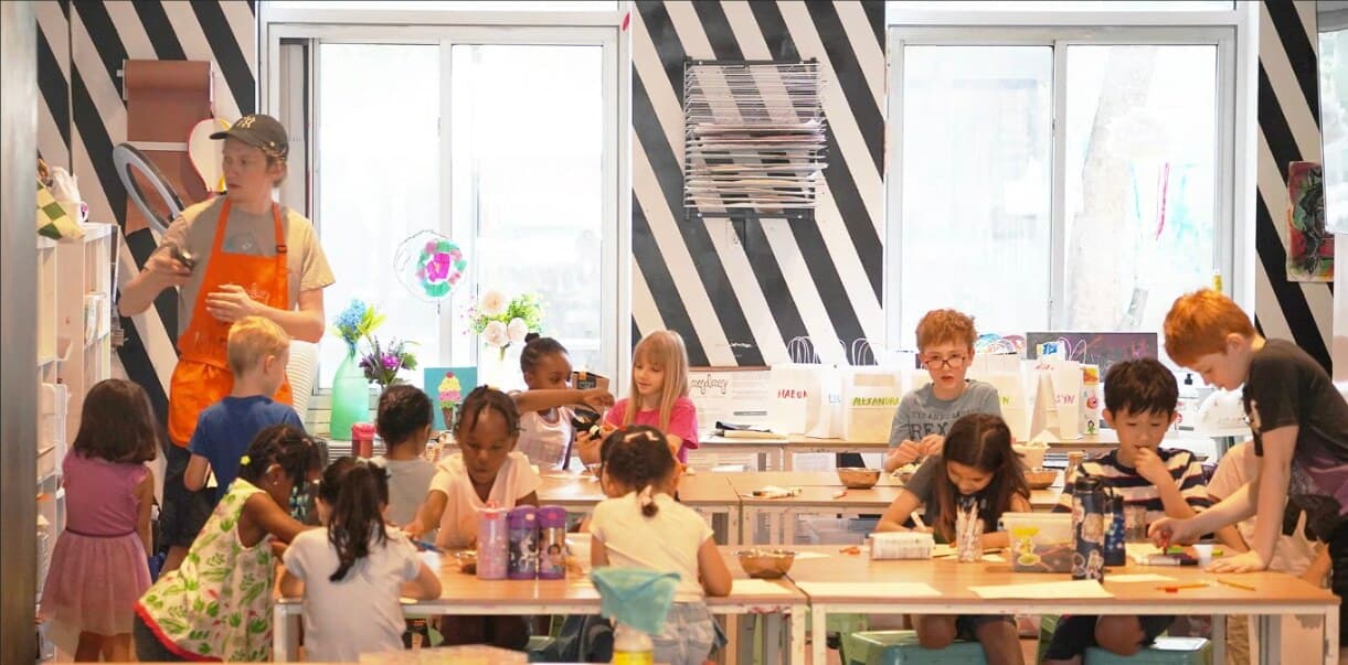 Kids working on art projects at long tables in the PLAYDAY studio, with a teacher in an orange apron